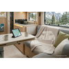Interieur van een camper: tafel met een tablet met kaart, twee mokken, bank met een deken met campersymbool, kussens, kitchenette en raam met uitzicht op bergen en bos