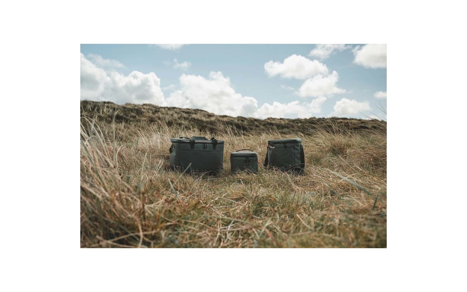 Tres cajas rectangulares verdes sobre hierba seca frente a dunas de arena bajo un cielo nublado
