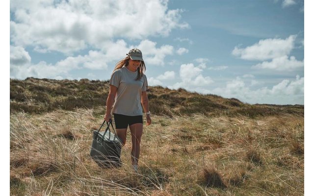 Persona caminando por dunas con hierba, viste camiseta, pantalones cortos y gorra de béisbol, lleva una bolsa grande; cielo nublado
