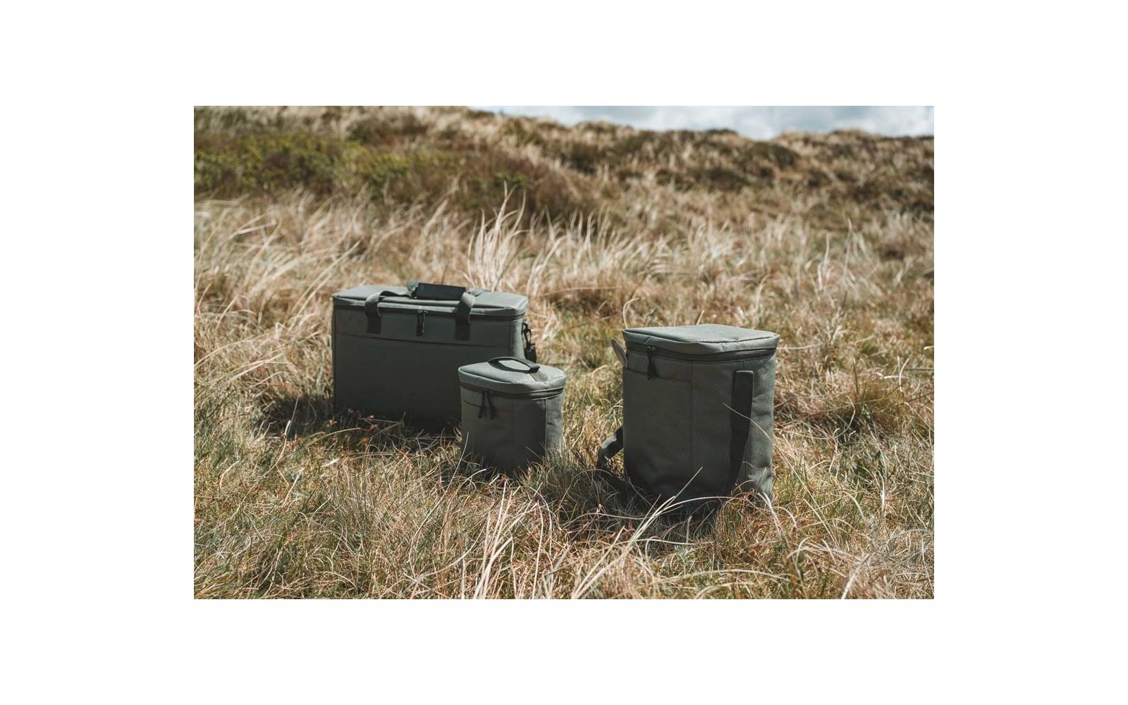 Tres neveras verde oliva de diferentes tamaños sobre hierba seca, dunas de arena al fondo