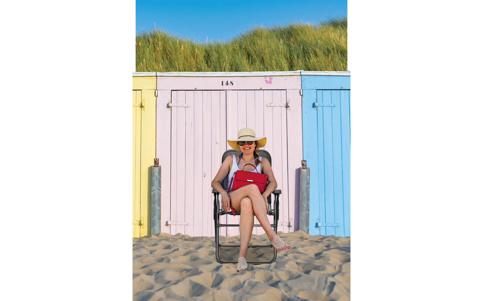 Persona sentada en una silla plegable frente a casetas de playa coloridas, con arena y dunas al fondo