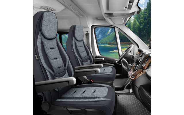 Innenansicht vorn eines Wohnmobils mit zwei grauen Sitzen, Lenkrad links, Armlehnen und Armaturenbrett; durch das Fenster ein See, Berge und Nadelwald