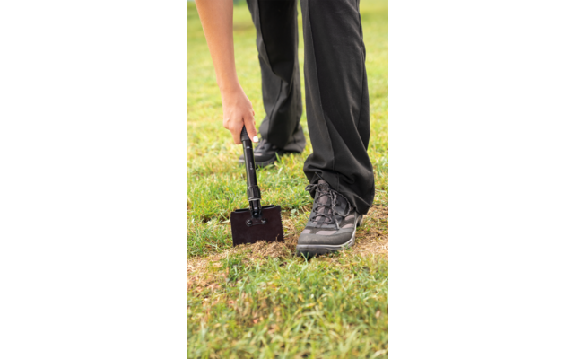 Unterkörper einer Person in schwarzen Hosen und grauen Schuhen, auf Rasen stehend, hält eine zusammenklappbare Schaufel, deren Blatt den Boden berührt