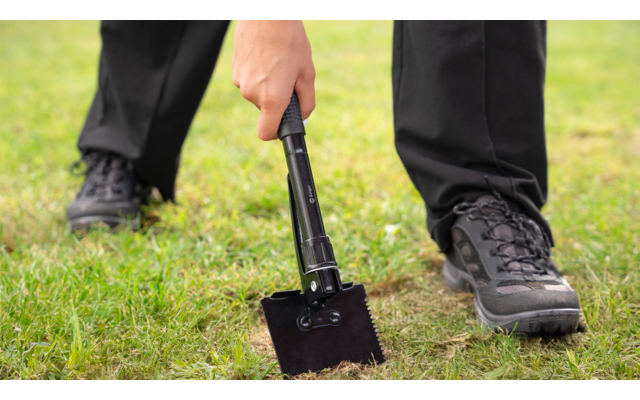 Person in schwarzer Hose und schwarzen Schuhen hält eine klappbare schwarze Schaufel, Schaufelblatt auf grasigem Boden