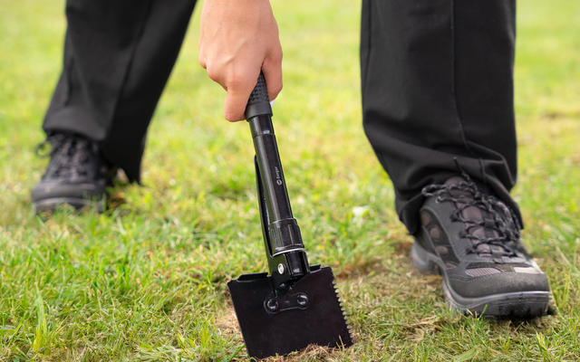 Personne en pantalon noir et chaussures noires tient une pelle noire pliante, la lame sur l'herbe