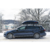 Station wagon blu scuro con box da tetto visto di lato su una strada innevata, alberi e un edificio innevati sullo sfondo