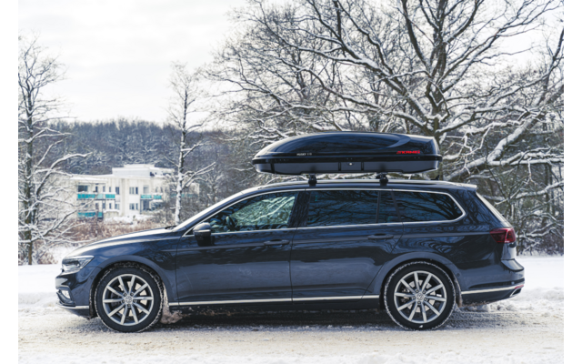 Station wagon blu scuro con box da tetto visto di lato su una strada innevata, alberi e un edificio innevati sullo sfondo
