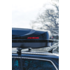 Box da tetto nero con logo rosso KAMEI montato sulle barre portapacchi di un'auto, alberi innevati sfocati sullo sfondo
