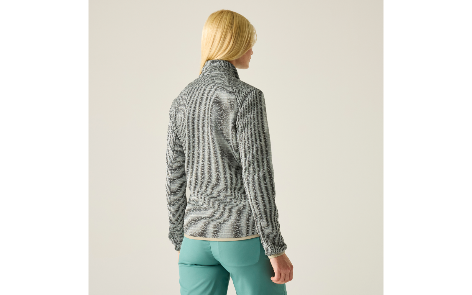 Person von hinten, trägt graue melierte Fleecejacke mit Stehkragen und türkisgrünen Hosen, lange blonde Haare, heller einfarbiger Hintergrund