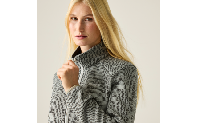 Blonde Frau trägt graue, gemusterte Jacke mit Reißverschluss, hält den Kragen mit der Hand, vor hellem Hintergrund