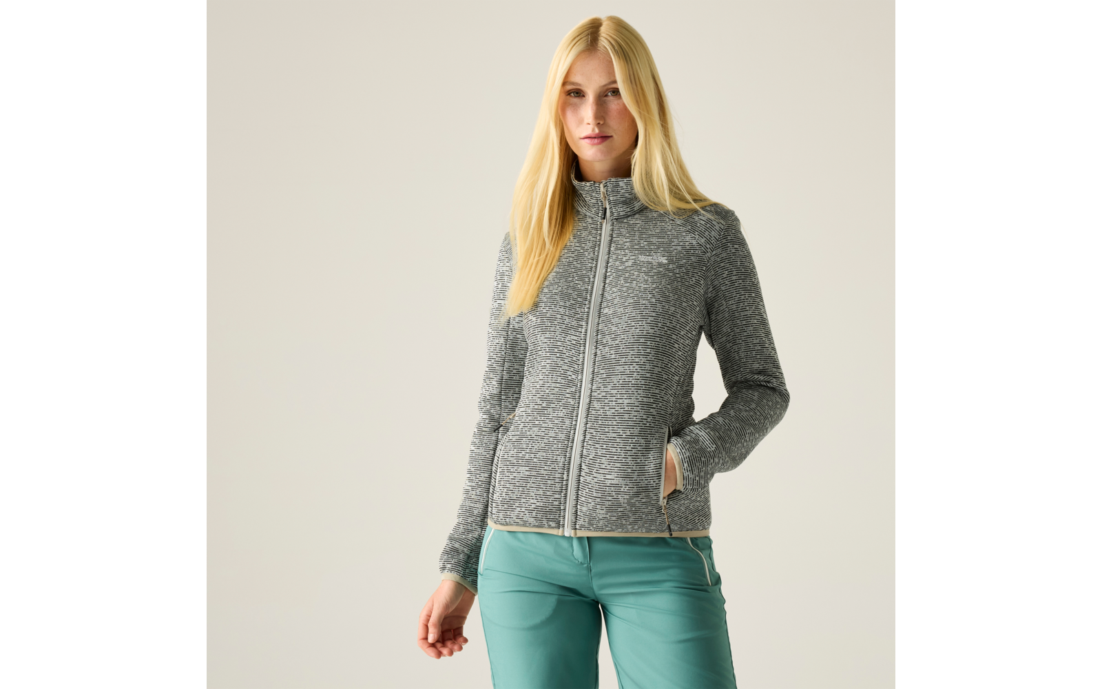 Person mit langen blonden Haaren, graue Jacke mit Reißverschluss und türkise Hose, eine Hand in der Hosentasche, einfarbiger heller Hintergrund