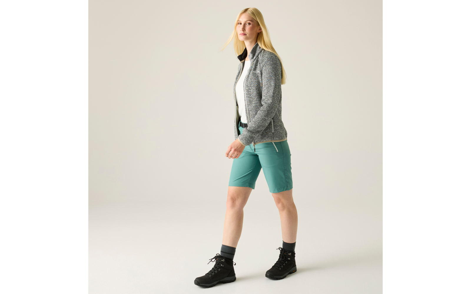 Person mit langen blonden Haaren, grauer Jacke, weißem T‑Shirt, grünen knielangen Shorts und schwarzen Wanderschuhen, geht vor hellem Hintergrund