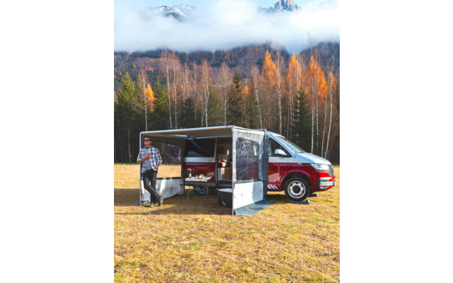 Person mit Sonnenbrille und kariertem Hemd steht neben rotem-weißen Van mit ausgefahrener Markise auf einer Wiese; im Hintergrund Bäume mit orangefarbenen Blättern und nebelverhangene Berge