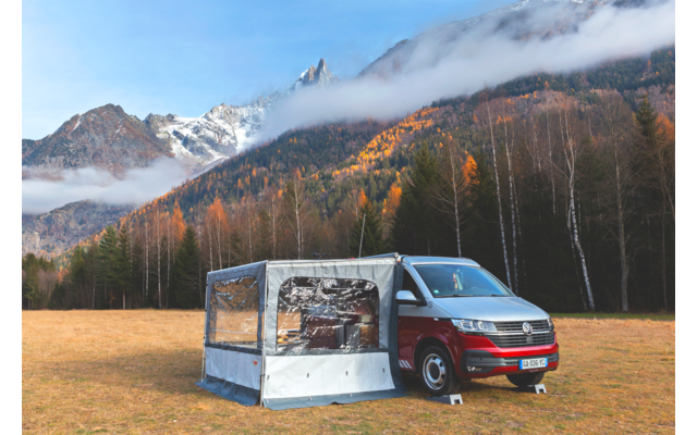Rotes Wohnmobil mit angebautem grauem Vorzelt auf einer Wiese; im Hintergrund herbstlich gefärbte Bäume, schneebedeckte Berge und niedrige Wolken