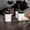 Drei weiße Behälter mit schwarzen Deckeln auf einer Arbeitsfläche, zwei Gläser Rotwein, eine Schale mit roten Trauben und eine Hand, die ein Etikett hält