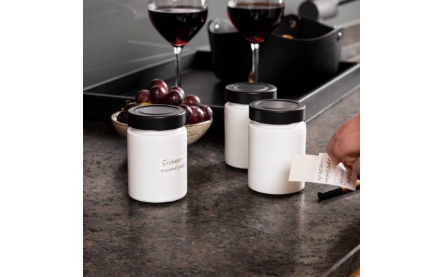 Drei weiße Behälter mit schwarzen Deckeln auf einer Arbeitsfläche, zwei Gläser Rotwein, eine Schale mit roten Trauben und eine Hand, die ein Etikett hält
