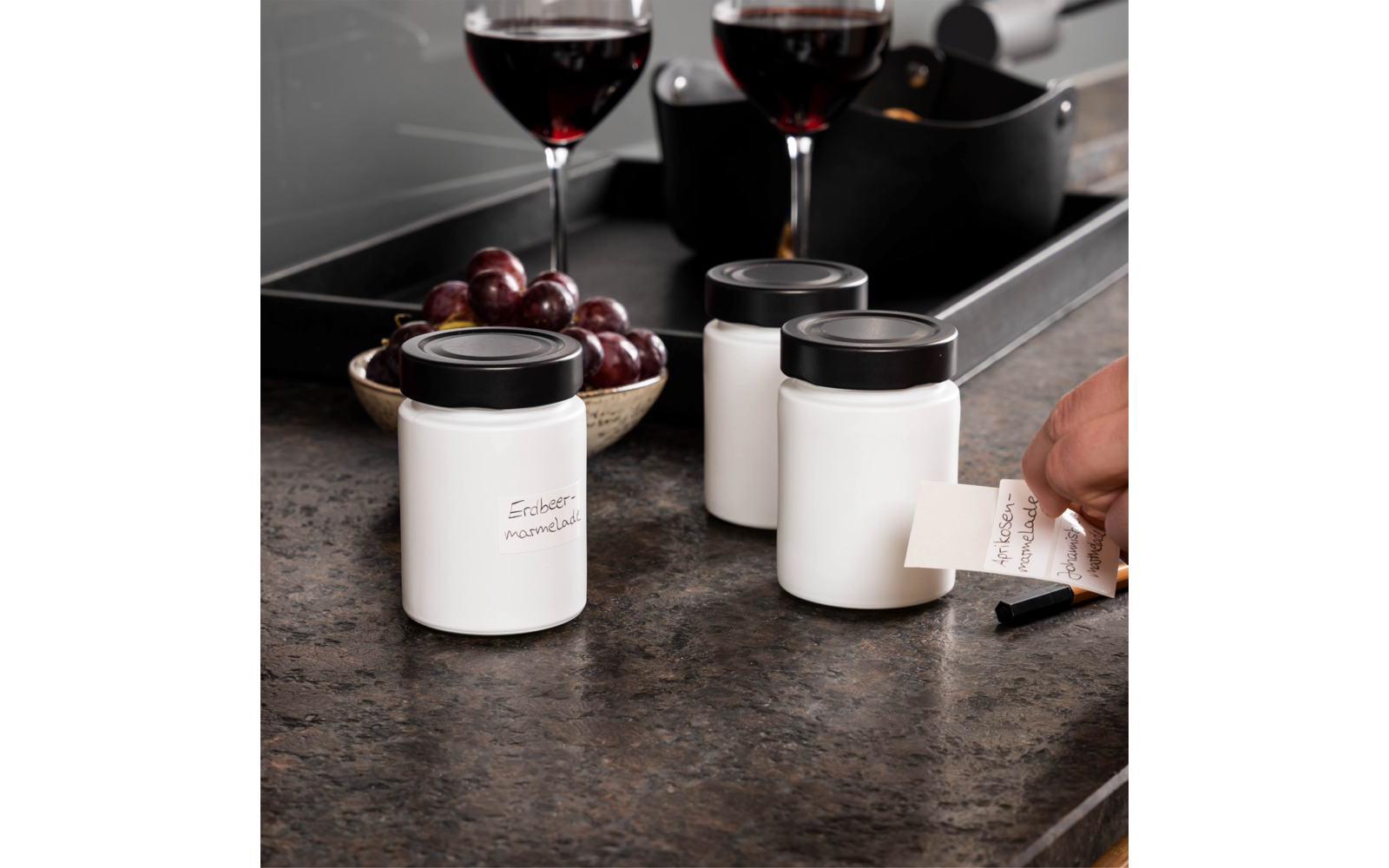 Drei weiße Behälter mit schwarzen Deckeln auf einer Arbeitsfläche, zwei Gläser Rotwein, eine Schale mit roten Trauben und eine Hand, die ein Etikett hält