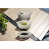 Three translucent, teardrop-shaped plastic bowls with lids side by side on light wooden decking; front bowl with black olives, middle with green grapes, back with green leaves and red pieces; potted plant at left, sunlight and shadows