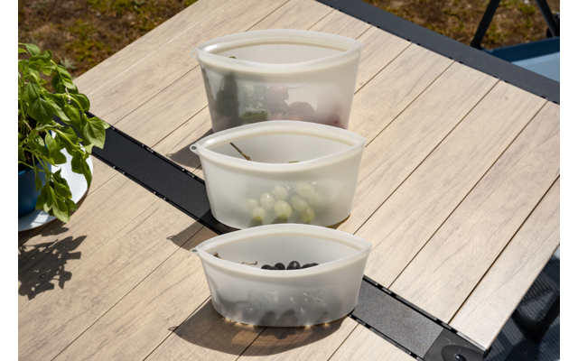 Three translucent plastic bowls in a row on a wooden outdoor table; front contains dark berries, middle contains green grapes, back contains red fruit