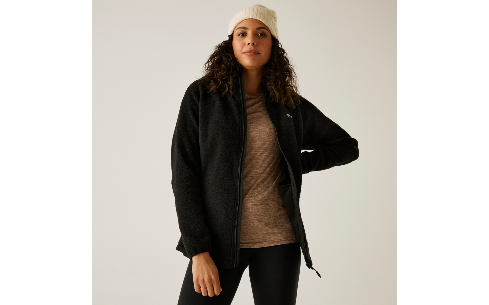 Frau mit lockigen Haaren trägt eine weiße Mütze, eine beige Strickjacke und einen schwarzen Reißverschluss-Pullover