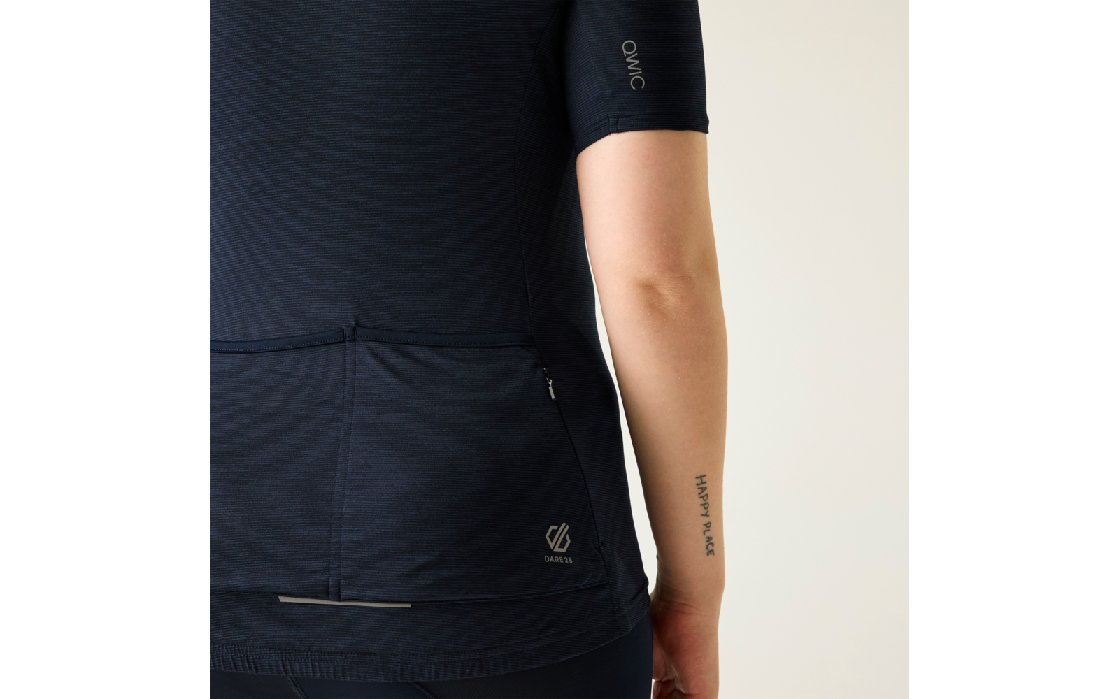 Rückenansicht einer Person von Taille bis Oberarm, trägt ein kurzärmeliges dunkelblaues Oberteil mit drei Rückentaschen und kleinem weißem Logo unten rechts, senkrechte Tätowierung mit Text am inneren Unterarm, heller Hintergrund