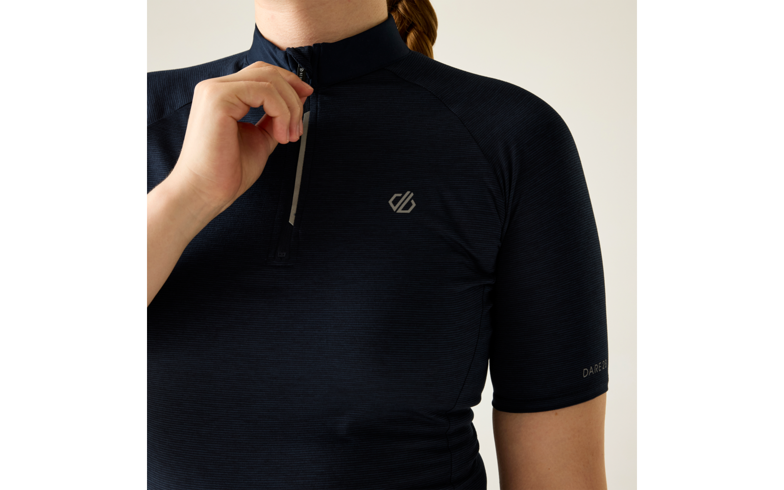 Oberkörper einer Person in dunkelblauem kurzärmeligen Oberteil mit kurzem Reißverschluss vorn, Hand hält den Reißverschluss, kleines Logo auf der Brust, Kinn und Nacken sichtbar, Haare zu einem Zopf gebunden, heller Hintergrund