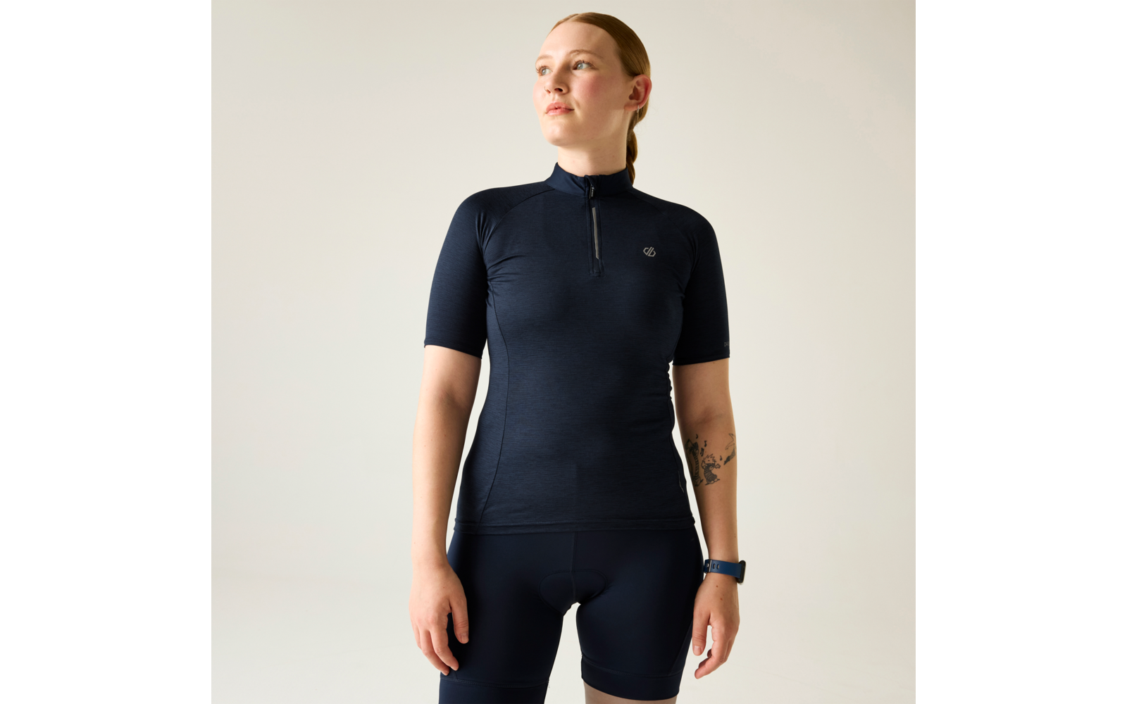 Person mit zusammengebundenem Haar, trägt dunkelblaues kurzärmeliges Trikot mit Reißverschluss und dunkelblaue Fahrradhose, Armbanduhr am Handgelenk, Tattoo am Unterarm sichtbar, Blick nach oben rechts