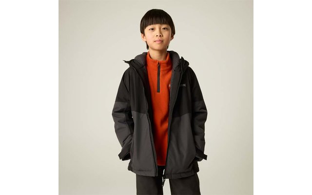 Junge steht frontal, trägt orangeen Reißverschluss-Pullover und schwarze-graue Jacke, Hände in den Taschen, hellbeiger Hintergrund