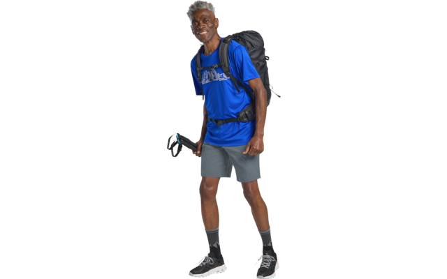 Persoon staat met rugzak en wandelstok, draagt blauw t‑shirt, grijze short, zwarte sokken en sportschoenen