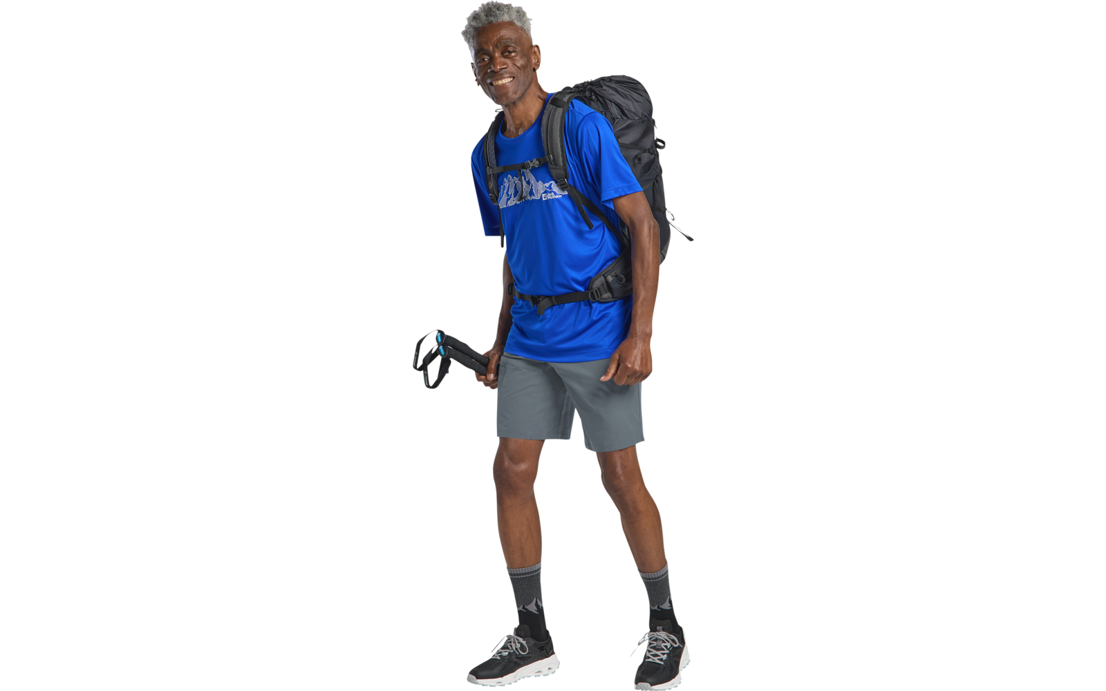 Persoon staat met rugzak en wandelstok, draagt blauw t‑shirt, grijze short, zwarte sokken en sportschoenen