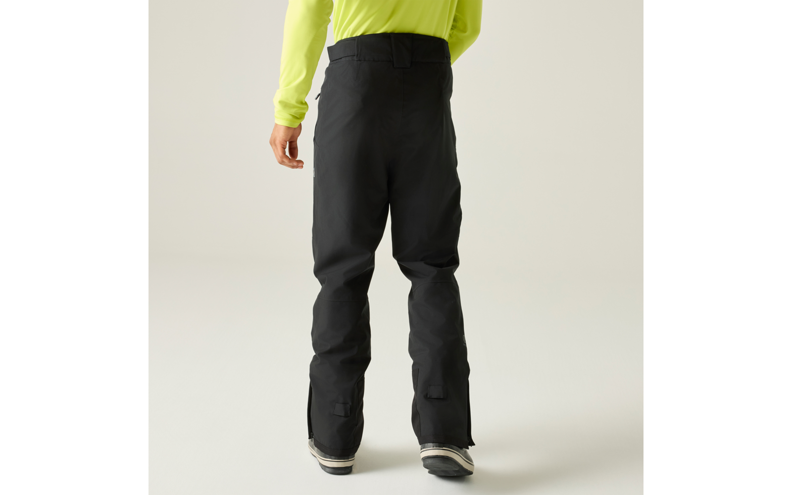 Vista trasera de una persona con camiseta de manga larga amarillo neón, pantalones negros anchos y botas negras sobre fondo claro
