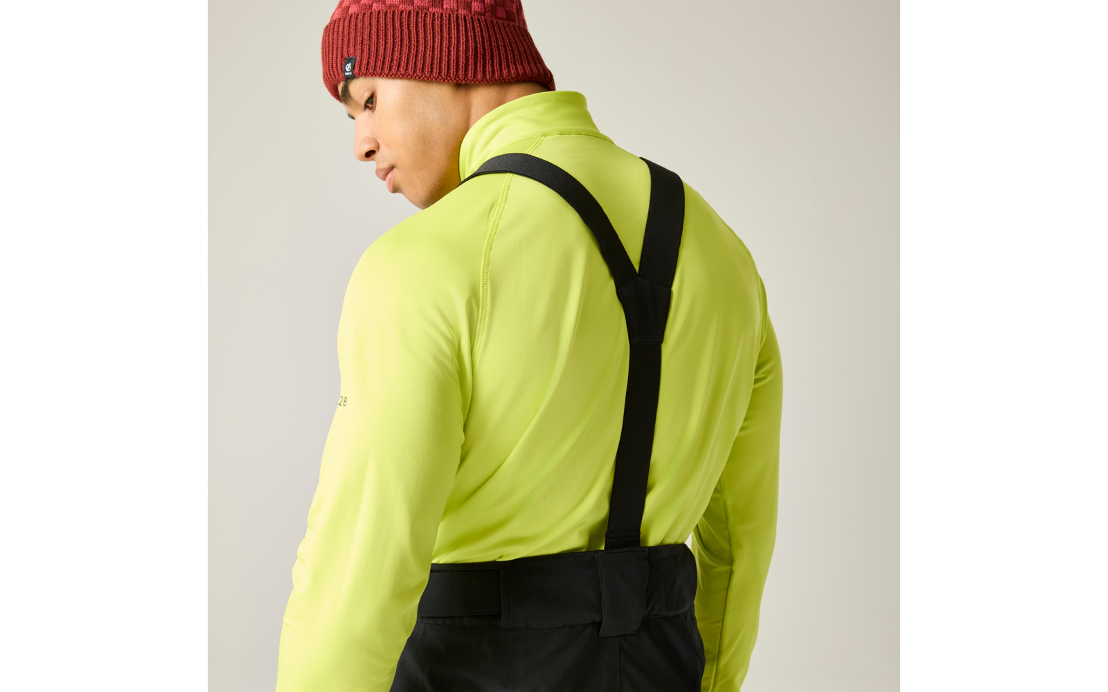 Persona vista de espaldas y de perfil, chaqueta amarillo neón, tirantes negros, gorro rojo de punto, fondo claro