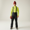 Persona de pie frente a la cámara sobre fondo claro, lleva camiseta de manga larga amarillo neón, pantalón negro con tirantes, gorro rojo de punto, sostiene guantes rojos, botas negras
