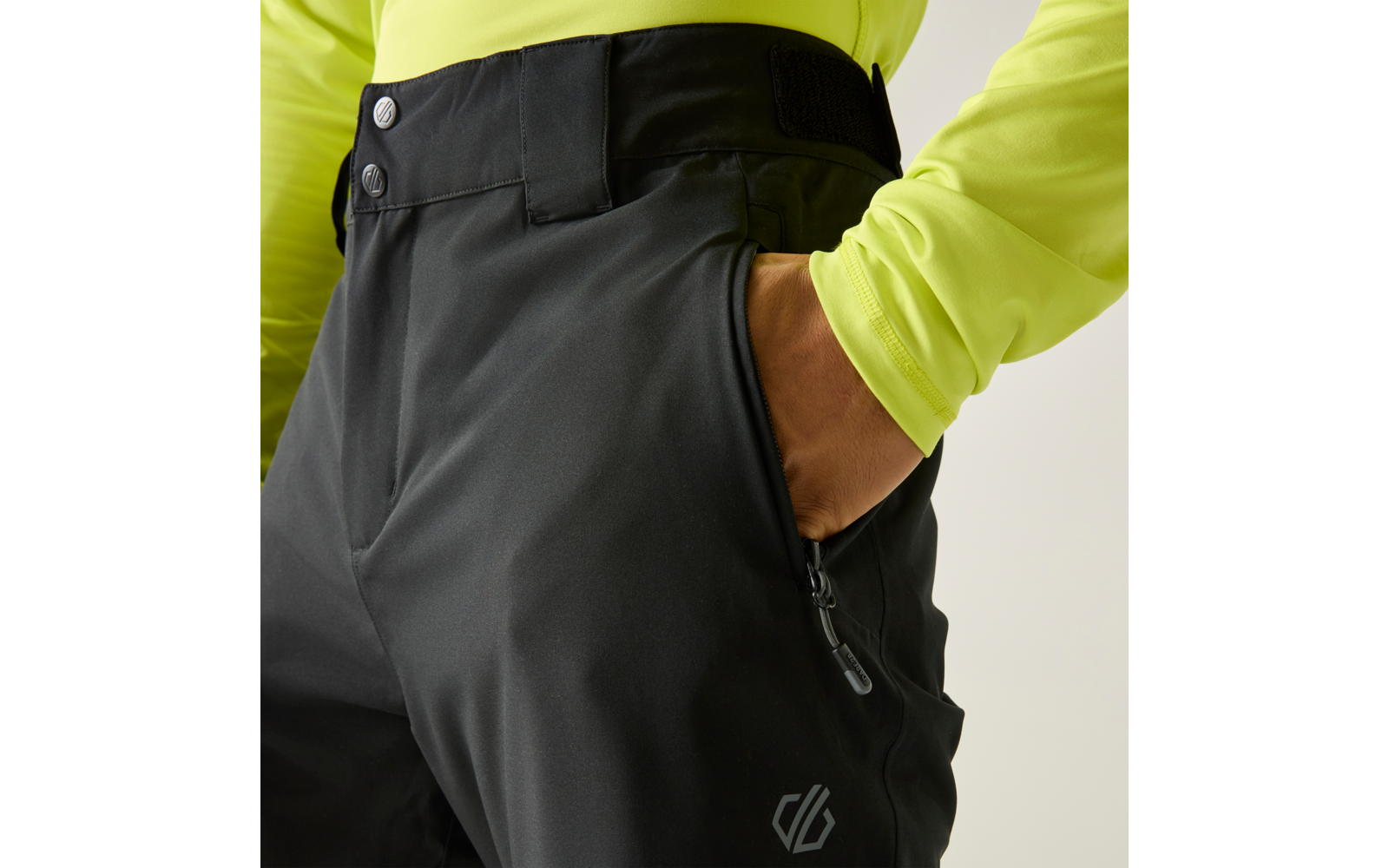 Mano en el bolsillo lateral de unos pantalones negros; cremallera, botones y velcro visibles en la cintura; encima, una camiseta de manga larga amarillo neón