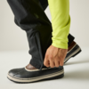 Mano cerrando la cremallera en la parte inferior de un pantalón negro; manga verde claro visible; zapato marrón con recubrimiento de goma negro y suela blanca