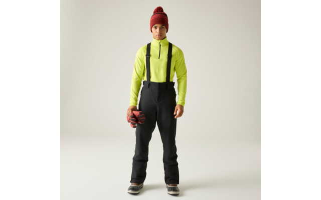 Persona de pie frente a la cámara sobre fondo claro, lleva camiseta de manga larga amarillo neón, pantalón negro con tirantes, gorro rojo de punto, sostiene guantes rojos, botas negras