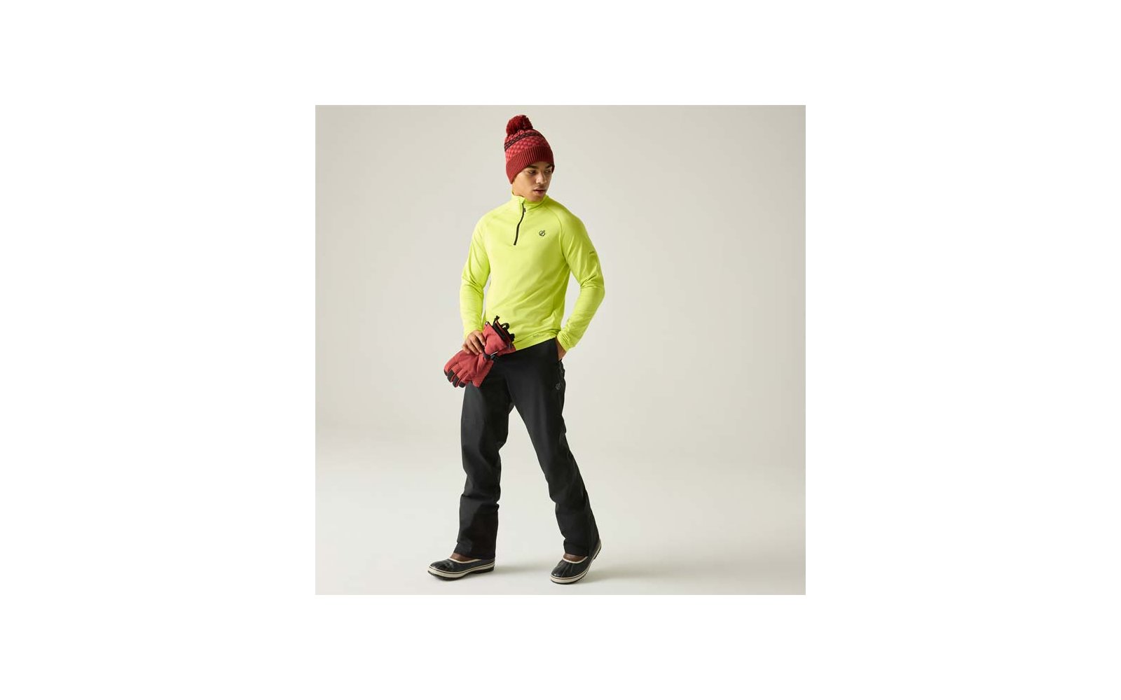 Persona con prenda superior verde neón con media cremallera, pantalones negros, gorro rojo de punto, guantes rojos en la mano, de pie sobre fondo claro