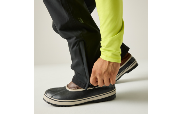 Mano cerrando la cremallera en la parte inferior de un pantalón negro; manga verde claro visible; zapato marrón con recubrimiento de goma negro y suela blanca