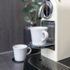 Weiße Espressomaschine auf grauer Arbeitsplatte, weiße Tasse auf dem Tropfblech unter der Auslaufdüse, weitere kleine weiße Tasse auf einem Untersetzer, dunkle Vase mit Eukalyptuszweigen im Hintergrund