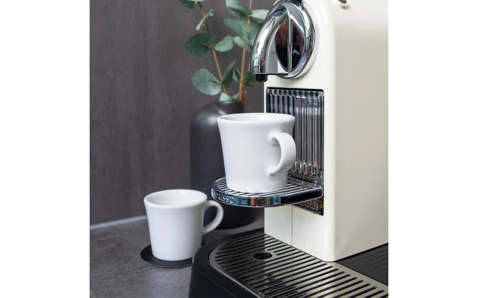 Weiße Espressomaschine auf grauer Arbeitsplatte, weiße Tasse auf dem Tropfblech unter der Auslaufdüse, weitere kleine weiße Tasse auf einem Untersetzer, dunkle Vase mit Eukalyptuszweigen im Hintergrund