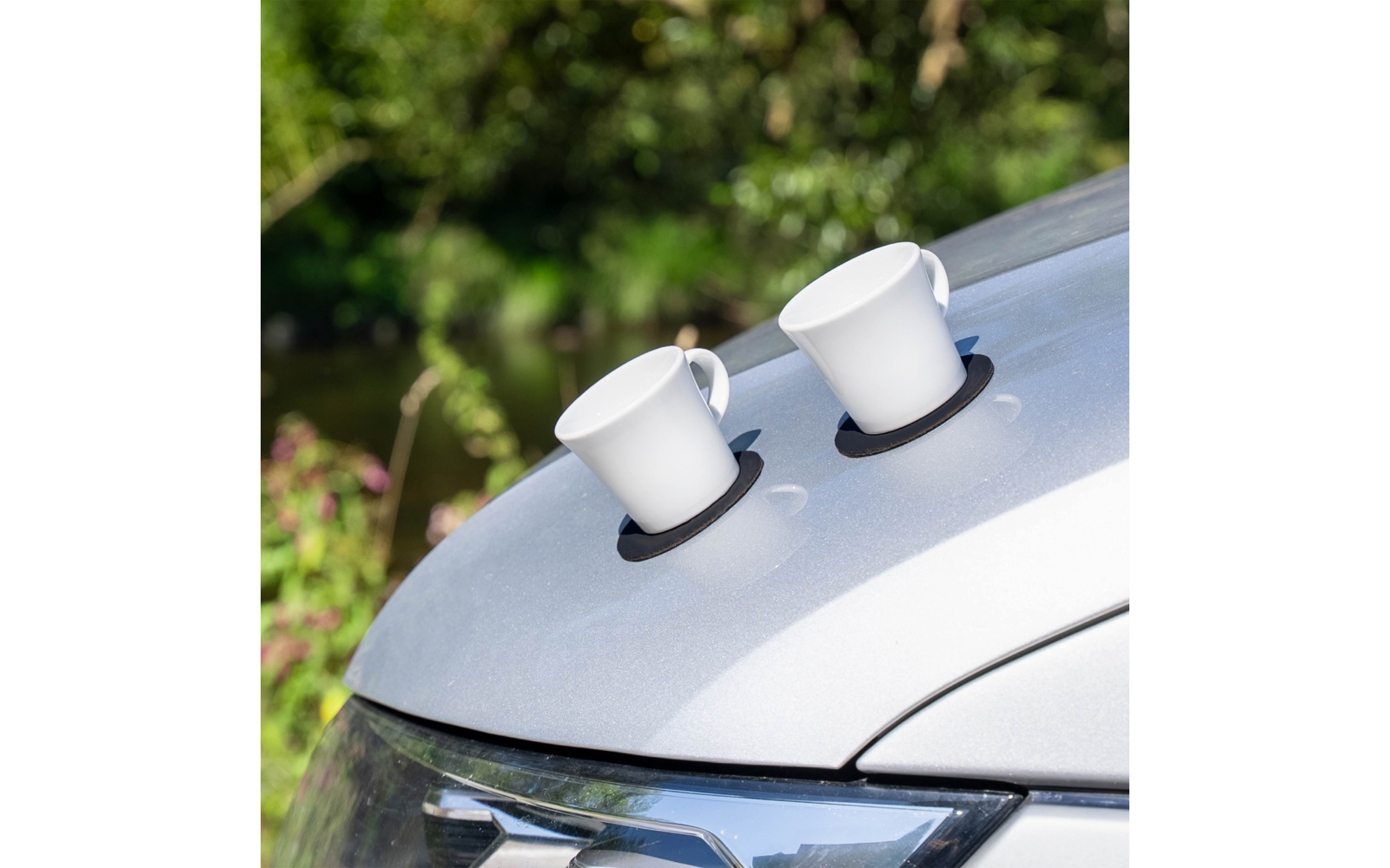 Zwei weiße Tassen auf schwarzen Untersetzern auf der Motorhaube eines silbernen Autos, unscharfer grüner Hintergrund