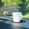 Weiße Tasse mit Henkel auf dunklem Untersetzer auf dem Armaturenbrett, im Hintergrund unscharfer Fluss und grünes Ufer
