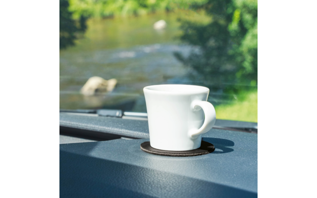 Weiße Tasse mit Henkel auf dunklem Untersetzer auf dem Armaturenbrett, im Hintergrund unscharfer Fluss und grünes Ufer
