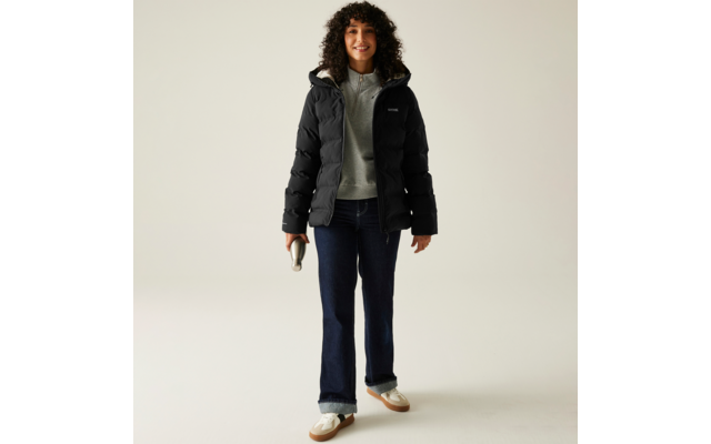 Frau trägt schwarze Jacke, grauen Pullover, blaue Hose und weiße Schuhe vor hellem Hintergrund