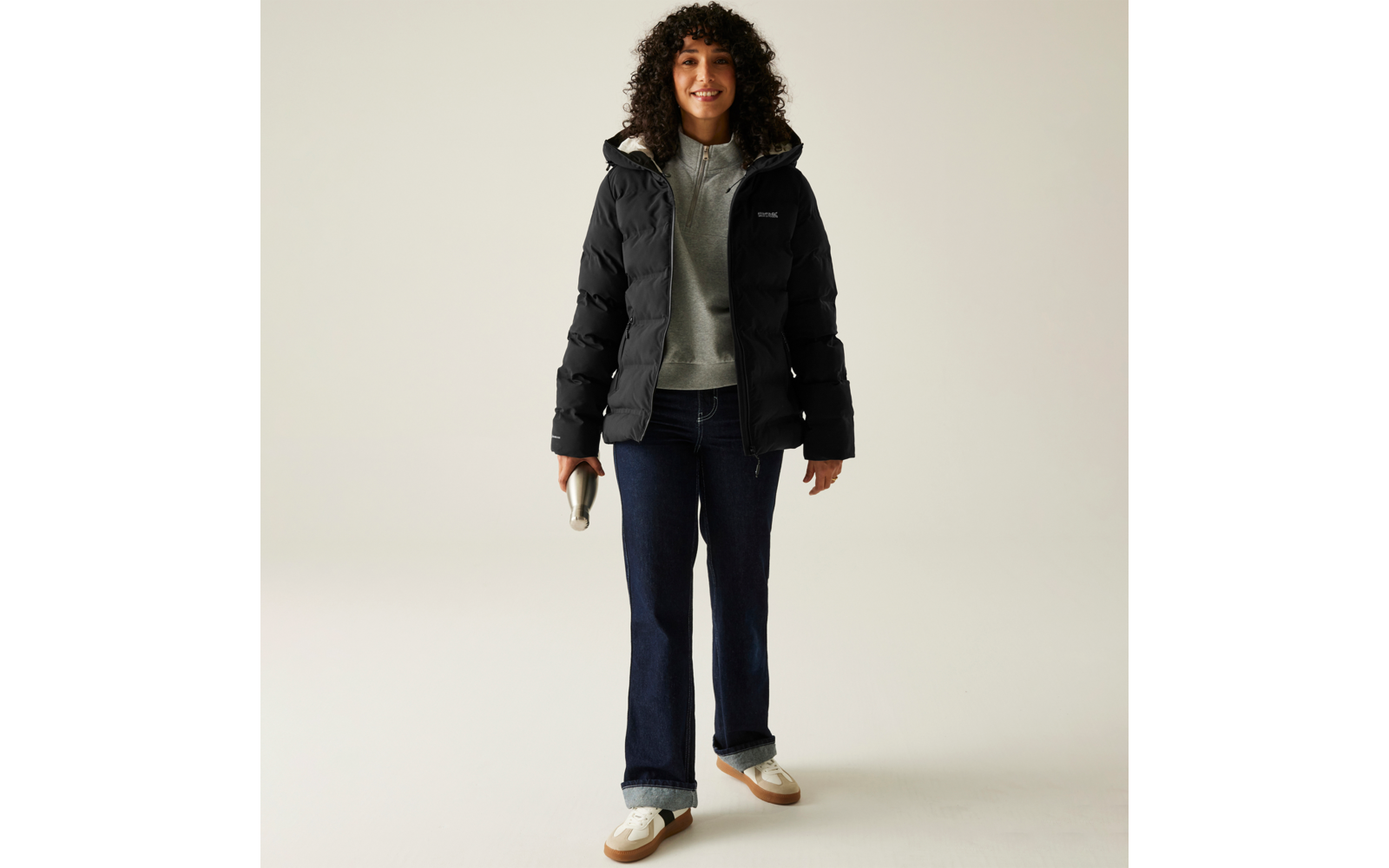 Frau trägt schwarze Jacke, grauen Pullover, blaue Hose und weiße Schuhe vor hellem Hintergrund
