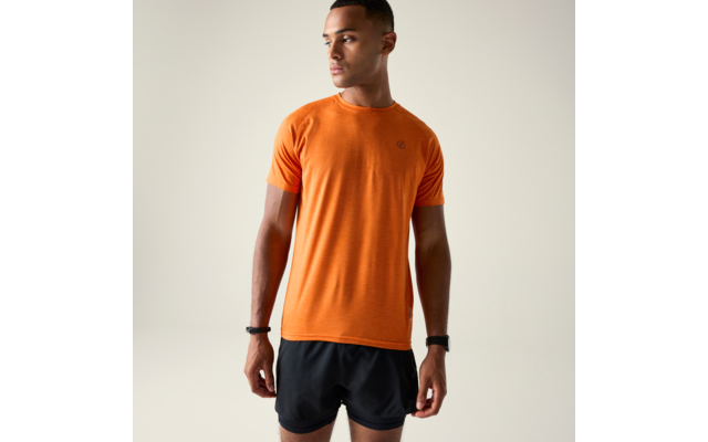 Personne debout portant un t‑shirt orange à manches courtes et un short noir devant un fond uni clair, regard tourné vers la droite