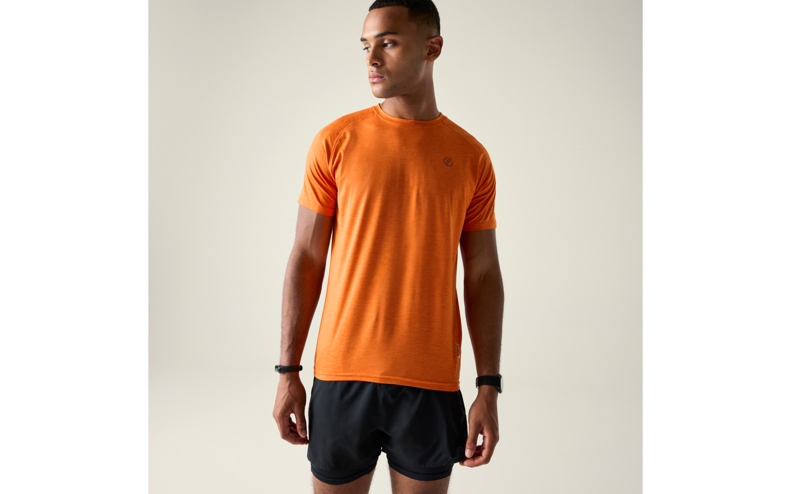 Personne debout portant un t‑shirt orange à manches courtes et un short noir devant un fond uni clair, regard tourné vers la droite