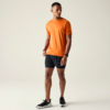 Homme aux cheveux courts debout, regardant vers la gauche, portant un t‑shirt orange, un short noir et des baskets noires, fond uni clair