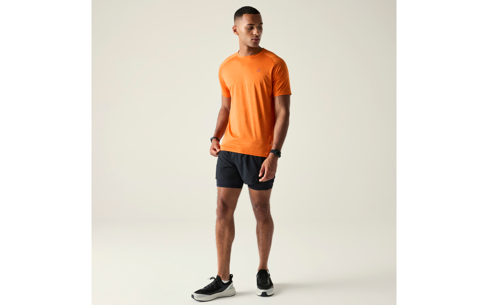Homme aux cheveux courts debout, regardant vers la gauche, portant un t‑shirt orange, un short noir et des baskets noires, fond uni clair