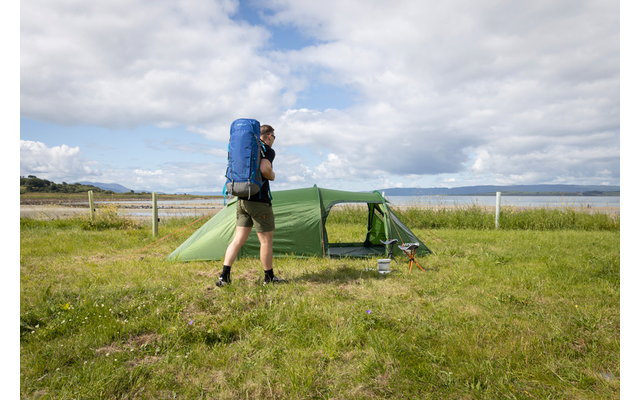 Person steht neben einem grünen Zelt auf einer Grasfläche, trägt blauen Rucksack, Shorts und Wanderschuhe; im Hintergrund Zaun, Wasser und bewölkter Himmel
